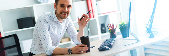 A young man is standing near a table in the office, holding a pencil and a glass of coffee. A young man works with documents and a computer.