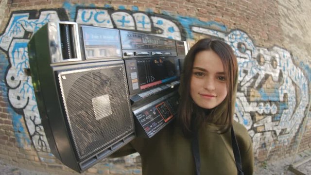 Cool stunning brunette with retro boombox looking into camera enjoying music, dancing. Portrait of cheerful girl in background of street art. Graffiti on wall. Outdoors.