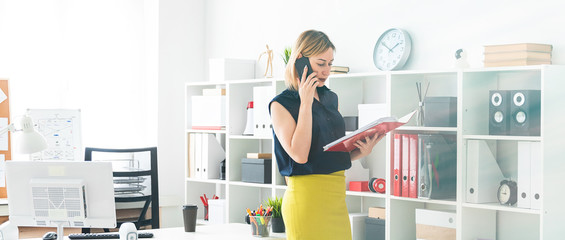 A young girl in the office talking on the phone and holding a folder with documents.