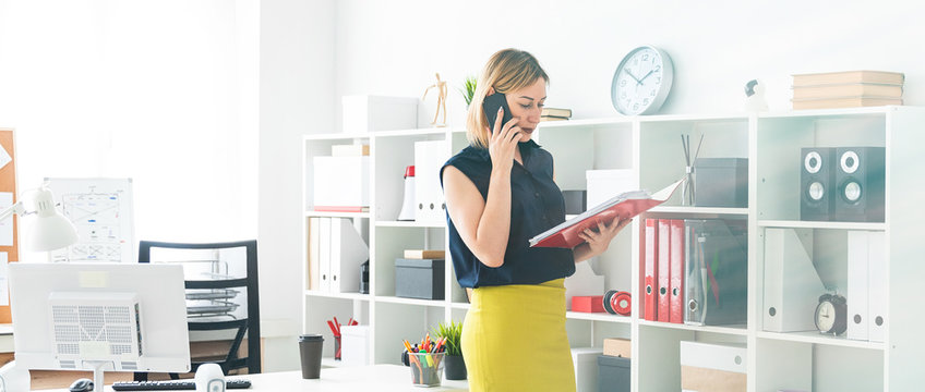 A young girl in the office talking on the phone and holding a folder with documents.