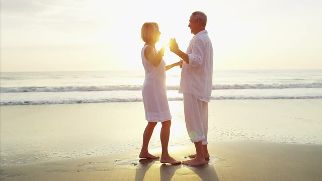 Mature Caucasian couple in white clothing dancing on the ocean beach at sunset