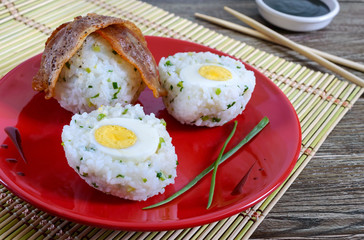 Boiled eggs wrapped in rice with green onions with slices of bacon on a red plate. Asian style breakfast.