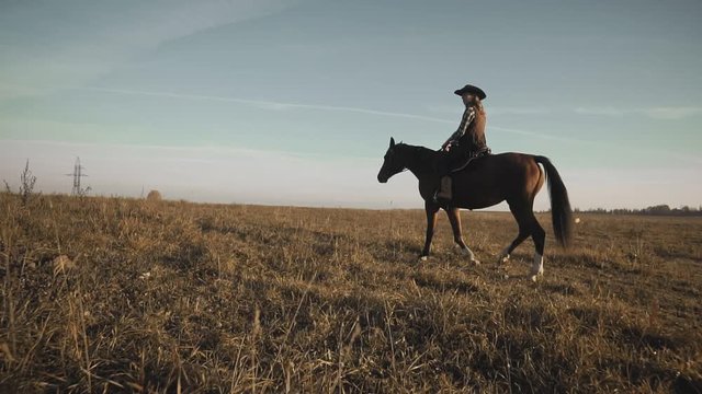 Beautiful cowgirl riding a horse in background sunrise in field. Young woman at brown horse in slow motion outdoors
