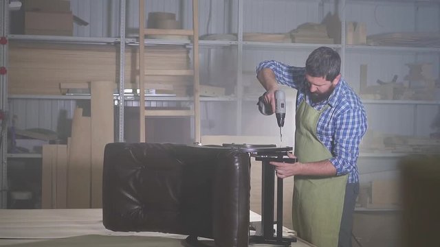 professional man with a beard in a shirt is engaged in underlaying assembling a furniture chair in a dark workshop