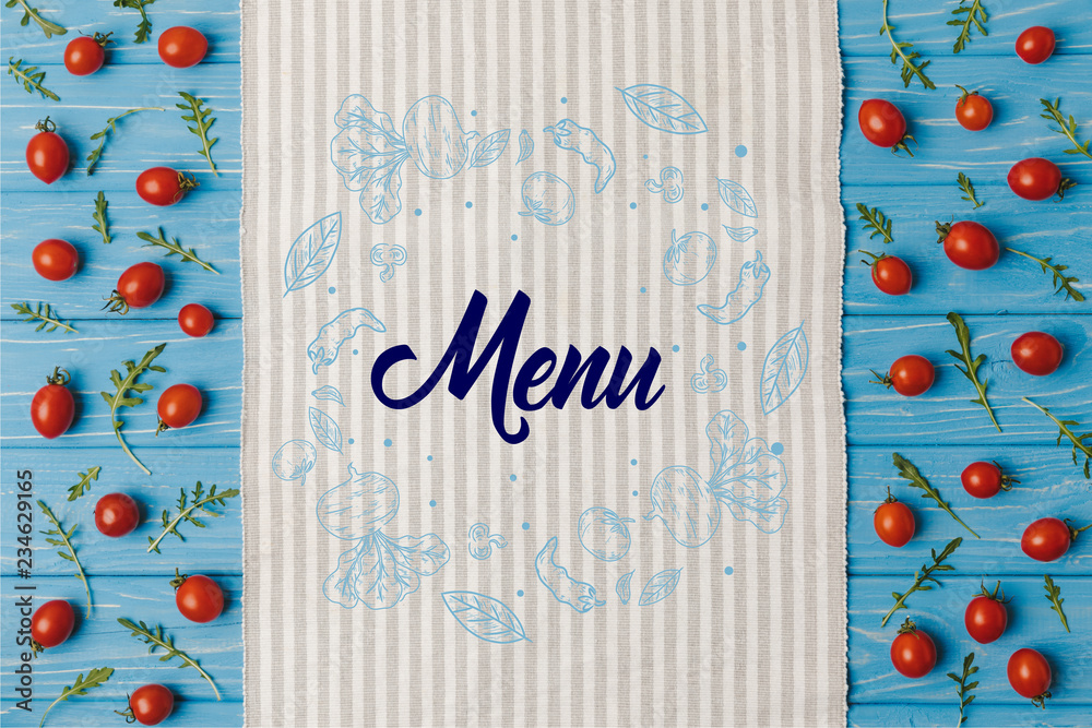 Wall mural top view of napkin and cherry tomatoes with arugula on blue table, menu lettering