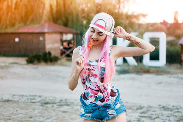 Happy girl on the beach wearing a cap, with pink hair licks a lollipop