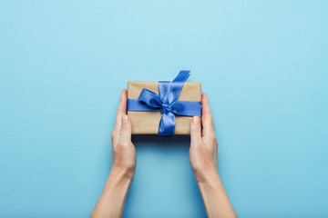 Female hands holding a gift with a blue ribbon on a blue background. Concept of a gift for the holidays, birthday, Christmas, wedding. Flat lay, top view