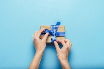 Female hands tying a bow on a gift box with a blue ribbon on a blue background. Concept of a gift for the holidays, birthday, Christmas, wedding. Flat lay, top view