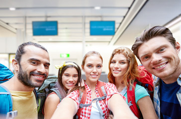 travel, tourism and people concept - group of smiling friends or tourists with backpacks taking selfie over airport terminal background