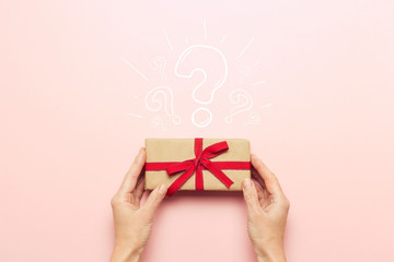 Female hands holding a gift box on a pink background with a question mark. Surprise, waiting for a gift for the holidays, birthday, christmas, wedding. Flat lay, top view