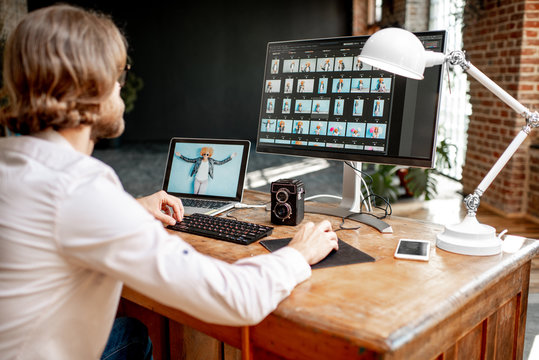 Young male photographer working with woman's portraits sitting at the working place with two computers in the studio