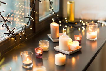 hygge, decoration and christmas concept - candles burning in lanterns and festive garland on window sill at home