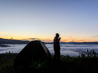 Silhouette of man standing with holding coffee cup near a camping tent at sunrise.