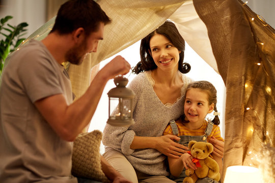 family, hygge and people concept - happy mother, father with lantern and little daughter with teddy bear playing in kids tent at night at home