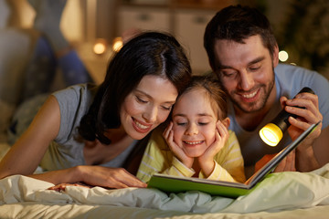 people and family concept - happy mother, father and little daughter reading book with torch light in bed at night at home