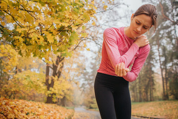 Sad and upset young female jogger stands in autumn park and hold hands on neck. She feels pain there. Young woman suffer.