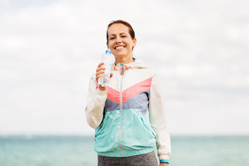 fitness, sport and healthy lifestyle concept - woman drinking water after exercising at seaside