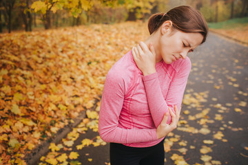 Sad and upset young woman suffer from pain in neck and elbow area. She keep seyes closed. Woman is in park alone.