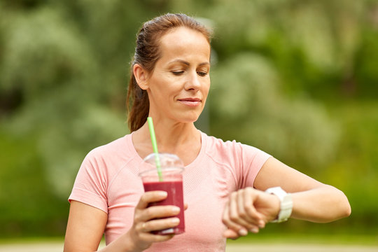 fitness, sport and healthy lifestyle concept - woman with takeaway smoothie drink or shake in plastic cup looking at smart watch in summer park