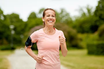 Foto auf Acrylglas Antireflex fitness, sport and healthy lifestyle concept - smiling woman with earphones wearing armband for smartphone, jogging at summer park and listening to music © Syda Productions