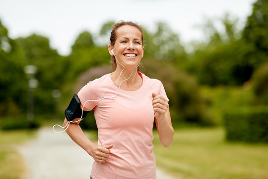 fitness, sport and healthy lifestyle concept - smiling woman with earphones wearing armband for smartphone, jogging at summer park and listening to music