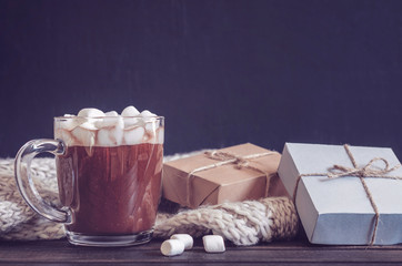 Hot cocoa with marshmallows in a glass cup surrounded by Christmas gifts and winter clothes, Christmas atmosphere and coziness