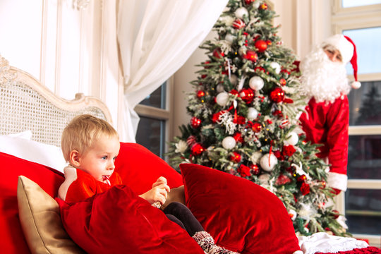 Little sad boy is sitting on a bed in a festive bedroom. The real Santa Claus is hiding from him behind the Christmas tree.