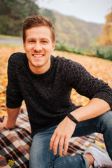 Portrait of young handsome man sitting on blanket and looks straight with smile. He leans on one hand. Guy poses.