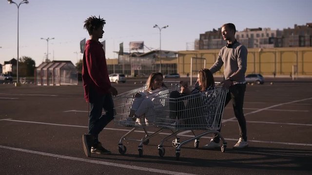 Multiethnic young people playing with shopping cart.s. Male friends standing near the shopping corts with their girlfriends. Young man explaining something to others. Outdoors on parking zone