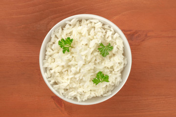 A bowl of cooked white jasmine rice, garnished with parsley leaves, shot from the top on a rustic wooden background with a place for text