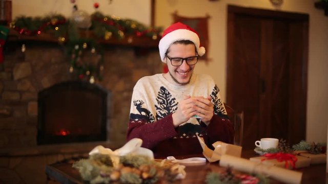 Happy man wearing santa hats opening Christmas gift box near fireplace with flashing garland. Smiling guy recieved a parcel with presents and enjoying unpacking package. Winter holidays concept.
