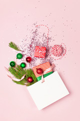 Flat lay top view White gift bag holographic glitter confetti red green christmas balls pine branches gift boxes on pink background. Greeting card Festive holiday decoration. Congratulations New Year