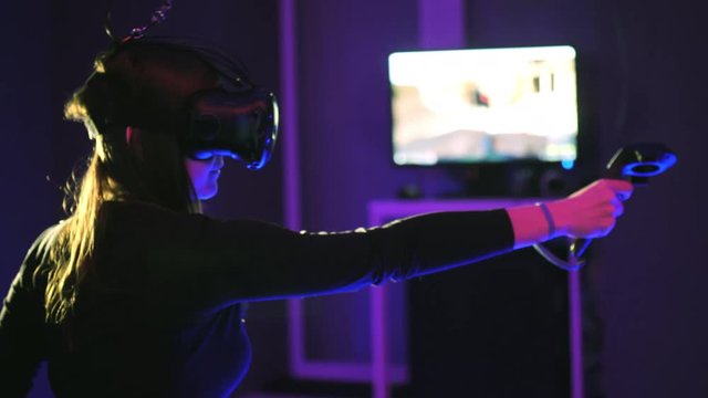 Woman play 3D virtual reality game. Shoots with controllers in a shooter VR game. Girl wearing VR headset in vr club. Monitor with the game in the background