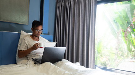 Asian business man have beard siting in the bed in the morning, Work on  notebook in his lap, at the same time drink coffee, Concept working on the bed.