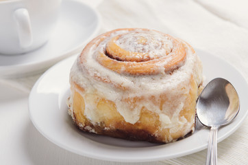 Hot cinnamon bun with sugar creamy icing on white plate. Sweet breakfast or snack with a cup of coffee.