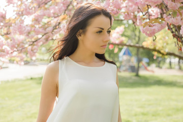 Attractive brunette woman in light dress looking at camera while posing near flowering tree