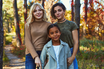 A beautiful couple of lesbian ladies posing in the autumn park with their adopted teenage daughter. The happy young family standing, looking at the camera, smiling. The girls wearing casual outfit.