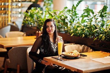 Pretty indian girl in black saree dress posed at restaurant, sitting at table with juice and salad.