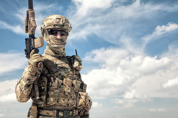Half-length portrait of airsoft game player in army camouflage uniform, tactical helmet, load carrier and face hidden behind mask, posing with firearm replica in hands, cloudy sky on background