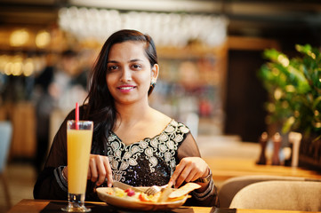 Pretty indian girl in black saree dress posed at restaurant, sitting at table with juice and salad.