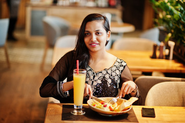 Pretty indian girl in black saree dress posed at restaurant, sitting at table with juice and salad.