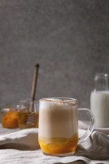 Glass of pumpkin layered spice latte with pumpkin puree, milk foam and cinnamon standing with ingredients in jars and decorative white pumpkins on crumpled table cloth with grey wall at background.
