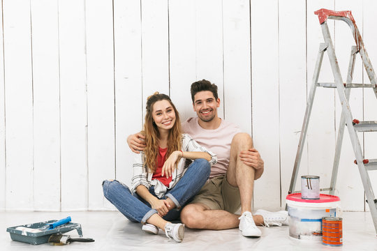 Photo of happy couple man and woman sitting on floor over white wall near ladder, while making renovation indoor