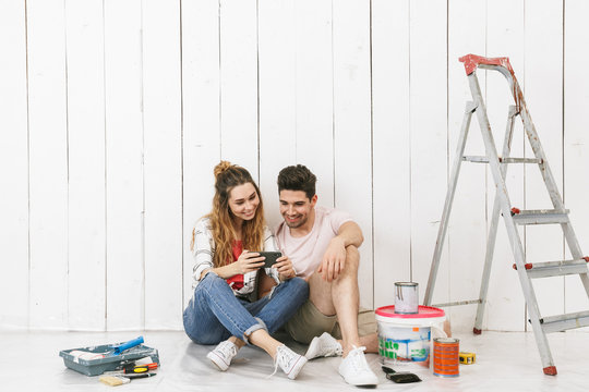 Photo of cute couple man and woman sitting on floor over white wall near ladder and using mobile phone, while making renovation indoor