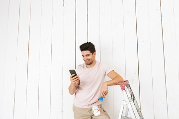 Photo of young man 20s using mobile phone, while painting white wall with paint roller and making...