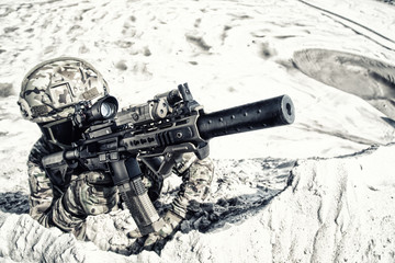 Airsoft player in military camouflage uniform, helmet and glasses lying in sand and aiming in...