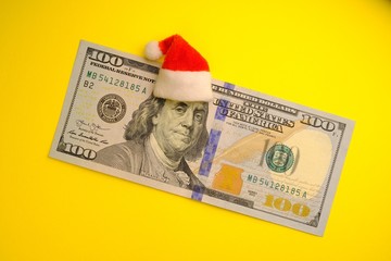 Franklin President in a red Santa Claus hat on a 100 US dollar bill on a bright yellow background. The concept of the cost of Christmas holidays, debt, profits or discounts for the new year.