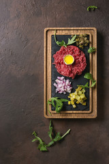 Beef tartare with quail egg in shell, cutting pickled cucumbers, capers, red onion, chives, arugula served on wooden black slate board with pepper mill on dark texture background. Flat lay, space