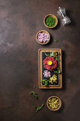 Obraz na płótnie Canvas Beef tartare with quail egg in shell, cutting pickled cucumbers, capers, red onion, chives, arugula served on wooden black slate board with pepper mill on dark texture background. Flat lay, space