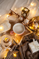 Fototapeta na wymiar Mug of hot cappuccino on a wooden tray is on the bed. Cozy decor. Breakfast. Mug, plaid, cotton, candle. Gift box and knitted mittens. Christmas lights. Holidays. Christmas. Autumn. Winter.